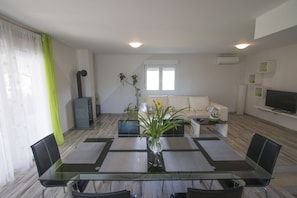 A modern living room with a glass dining table, black chairs, a white sofa, and a TV on the wall.
