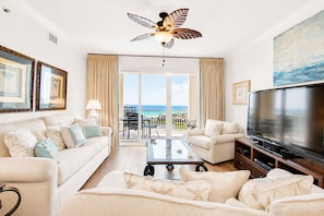 A spacious living room with a large TV, comfortable seating, a ceiling fan, and a balcony with a view of the ocean.