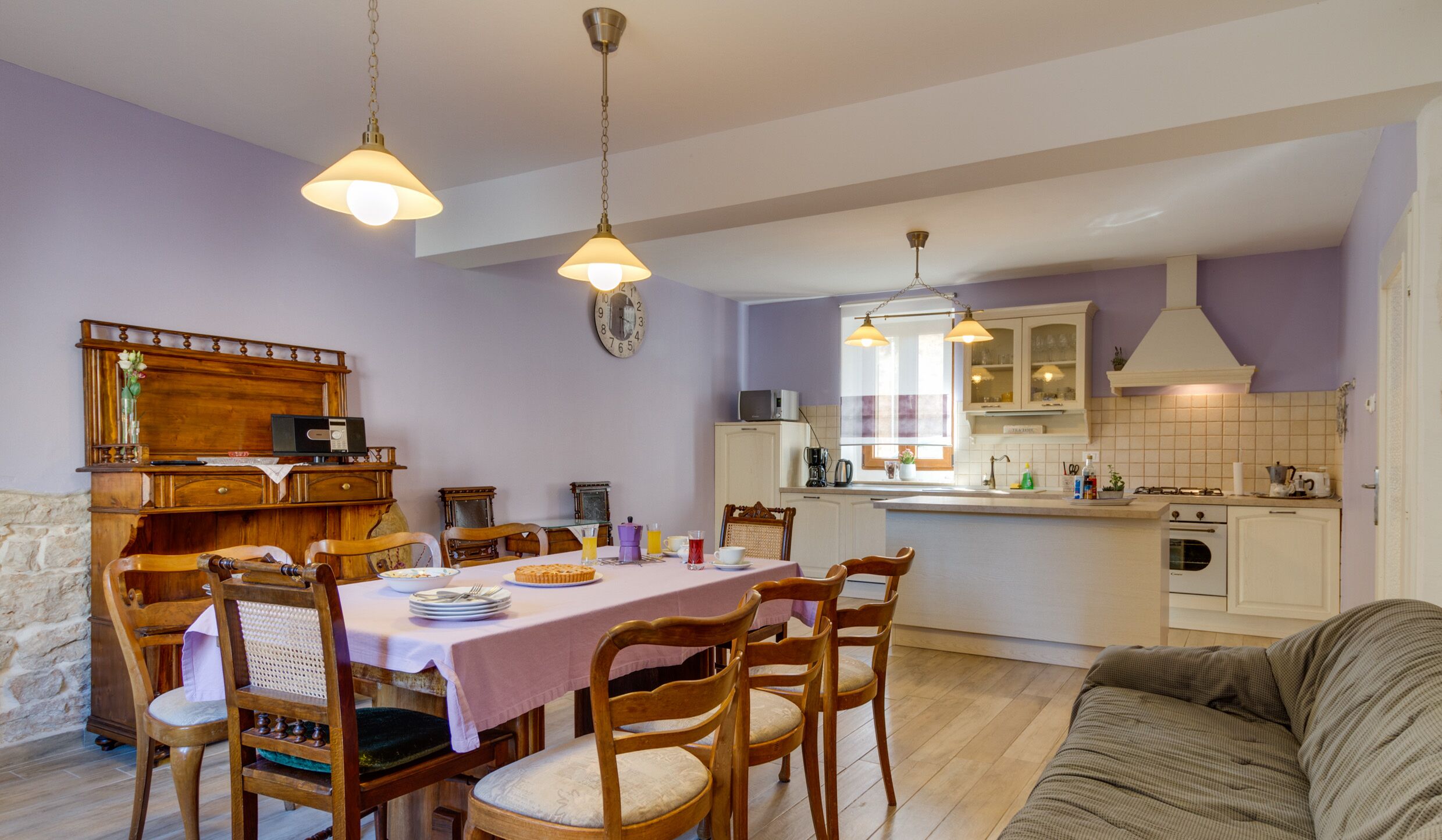 A dining area with a table set for four, wooden chairs, and pendant lights. There is a kitchen area with white cabinets and a stove.