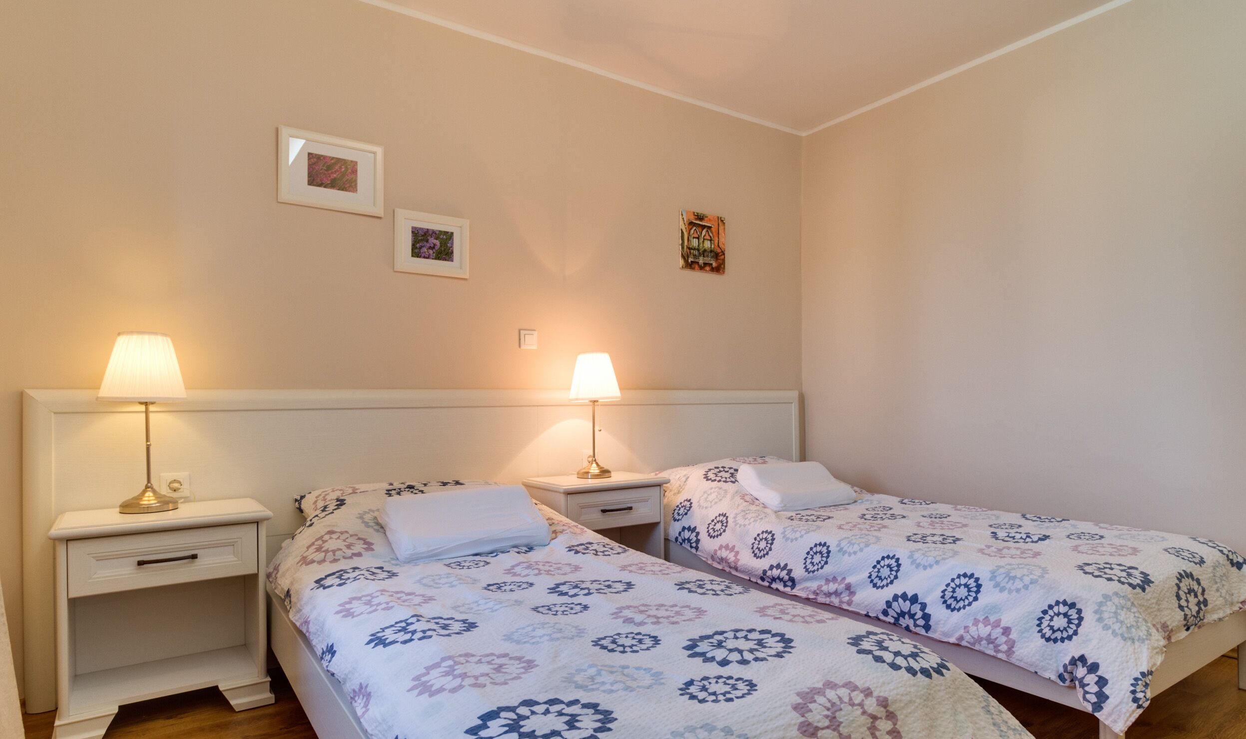 Habitación doble con dos camas individuales, cada una con mesita de noche y lámpara. Hay tres cuadros enmarcados en la pared.