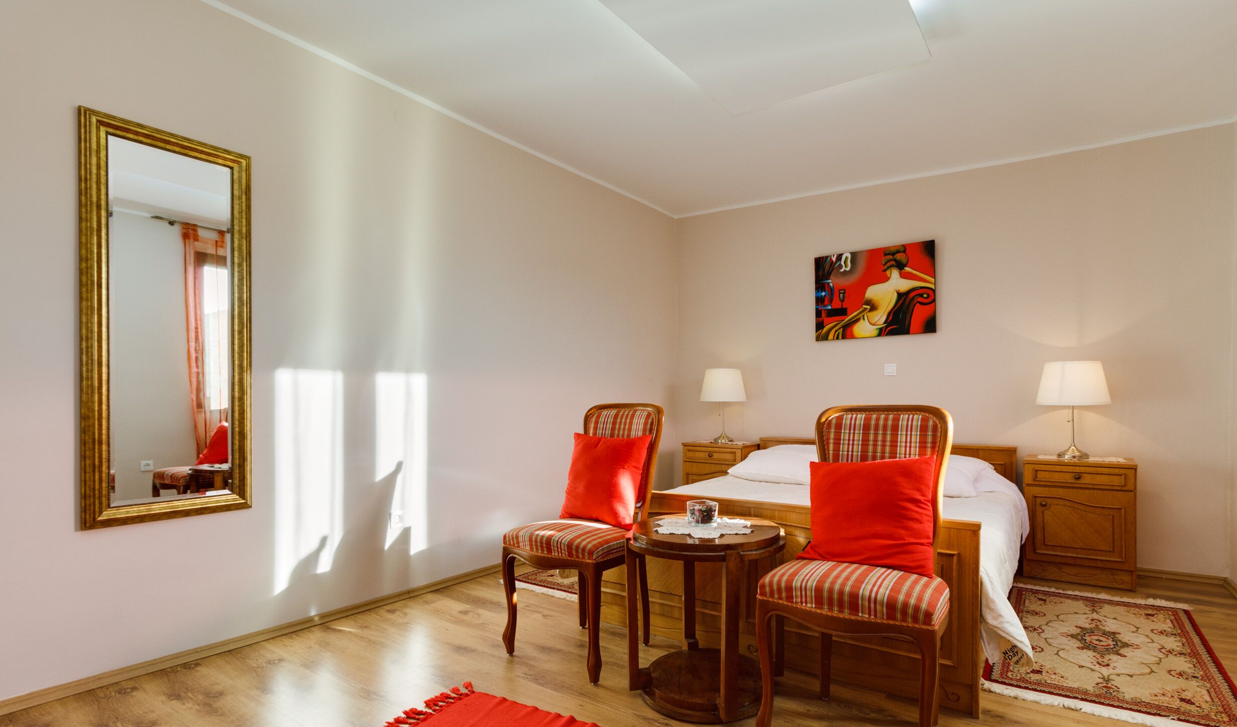 Una habitación de hotel con una cama, dos sillones, una mesa, una mesita de noche con lámparas, un cuadro y un espejo.