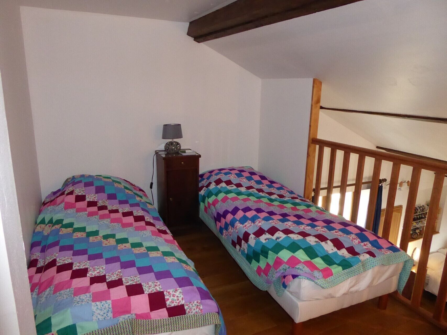A small, cozy room with two single beds, a nightstand with a lamp, and a wooden staircase.