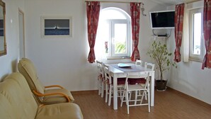 Een woonkamer met een beige bank, een eettafel met witte stoelen, een potplant en een aan de muur bevestigde televisie.