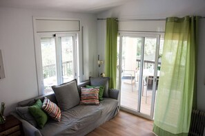 A living room with a grey sofa, green and striped pillows, a small table with a lamp, and a view through a glass door to an outdoor patio.