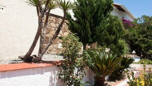 A two-story building with a white exterior and a red-tiled roof. There is a staircase leading up to the entrance, and various plants and shrubs are visible in the garden area. - Private villa with fantastic sea view! (Lloret de Mar)