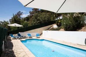 A swimming pool with blue water, surrounded by a white wall and a stone pathway. There are blue lounge chairs and a white umbrella providing shade.