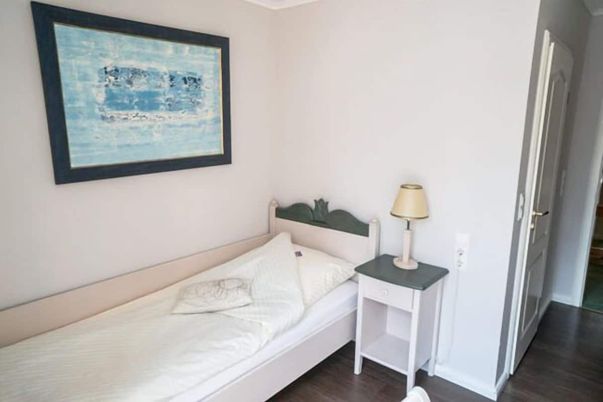 Ein Einzelbett mit weißem Kopfteil, ein Nachttisch mit Lampe und ein gerahmtes Kunstwerk an der Wand.