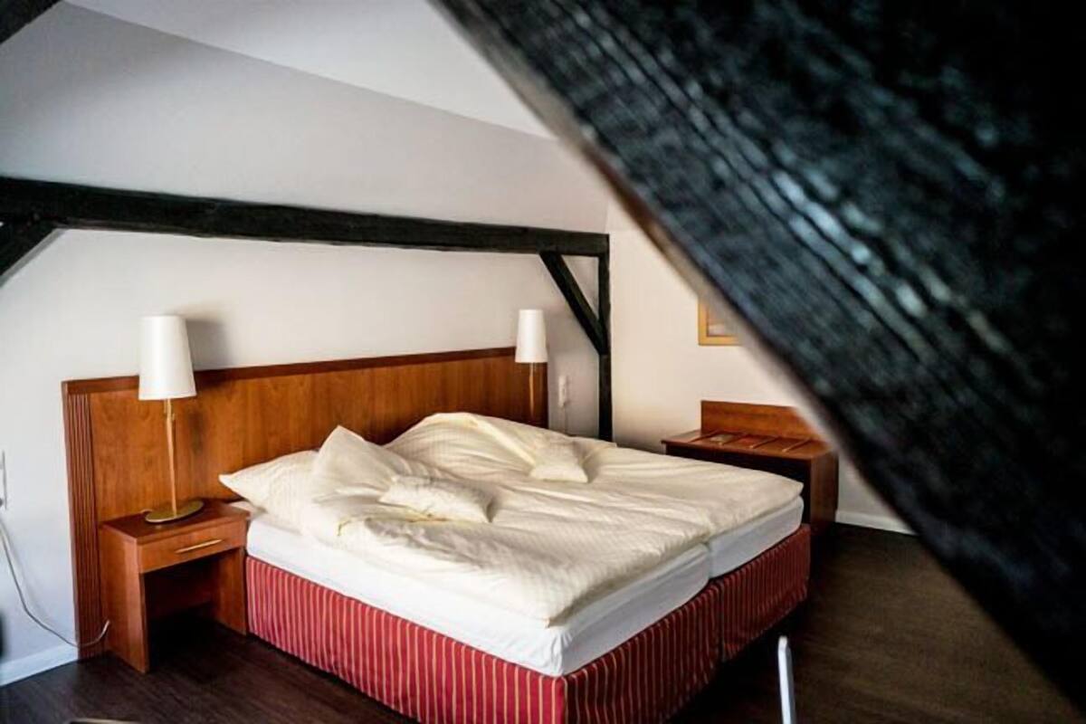 Ein Schlafzimmer mit einem hölzernen Kopfende, einem Bett mit weißen Leinentextilien, zwei Nachttischlampen und einem rot gestreiften Bettlaken.