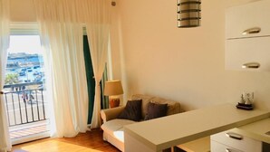 Une pièce avec un plancher en bois, un bureau blanc, un canapé et une lampe. Il y a une fenêtre avec des rideaux, et de là, on peut voir des bateaux à l’extérieur.