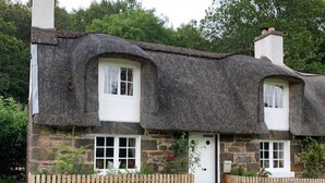 A traditional thatched-roof cottage with a stone facade, two windows, and a wooden fence.
