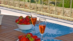 Ein Tisch mit zwei Schalen Erdbeeren und zwei Gläsern Orangensaft auf einer Holzterrasse, bedeckt mit einer blau gemusterten Tischdecke, mit Blick auf eine Küstenlandschaft.
