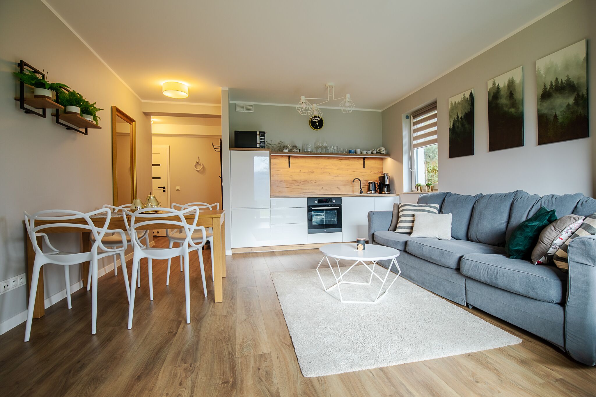 Una sala moderna con un sofá gris, sillas blancas y una mesa de comedor de madera. Se ve una cocina al fondo con armarios blancos y una encimera de madera.