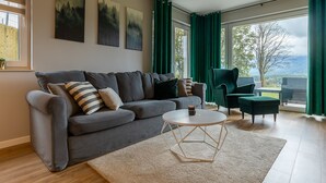 Een moderne woonkamer met een grijze bank, een ronde salontafel en uitzicht op het groen door grote ramen.