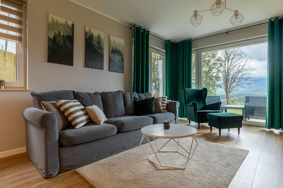 A modern living room with a grey sofa, a round coffee table, and a view of the outdoors through large windows.