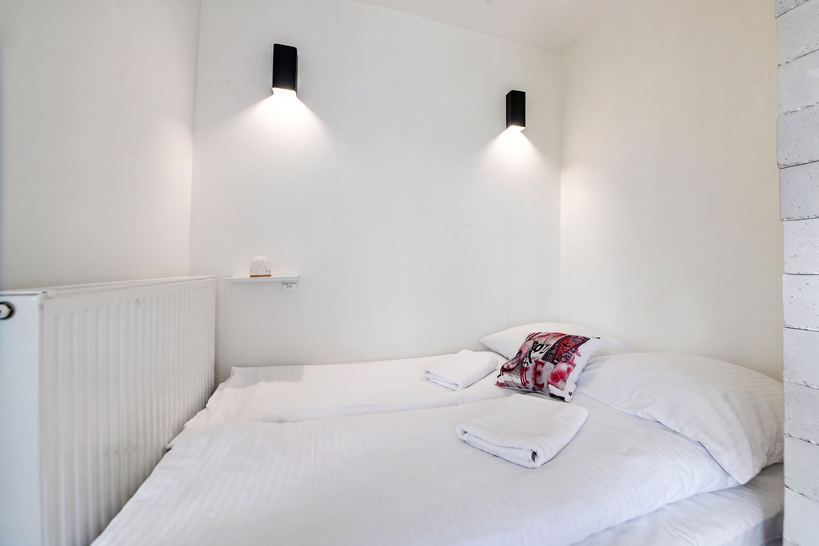 Una cama individual con sábanas blancas y una toalla doblada, una mesita de noche con una lámpara y un radiador.