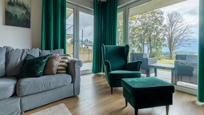 Een moderne woonkamer met een grijze bank, een groene fauteuil en een passende voetenbankje. Grote ramen bieden uitzicht op het omliggende landschap.