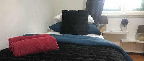 Una cama bien hecha con una colcha azul oscuro y toallas rojas, una mesita de noche blanca con una lámpara y una planta pequeña, y una ventana con cortinas.