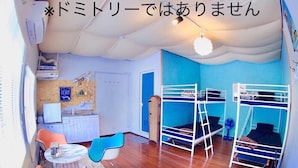 Una habitació amb llits superposats, una cuineta petita, una zona de menjador amb cadires i un ventilador de sostre.