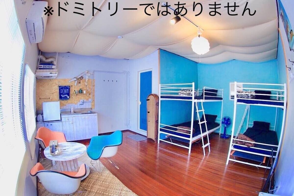 Quarto com beliches, uma kitchenette pequena, área de refeições com cadeiras e um ventilador de teto.