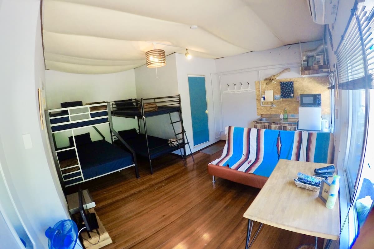 Quarto compacto com beliches, pequena cozinha e janela com cortinas.