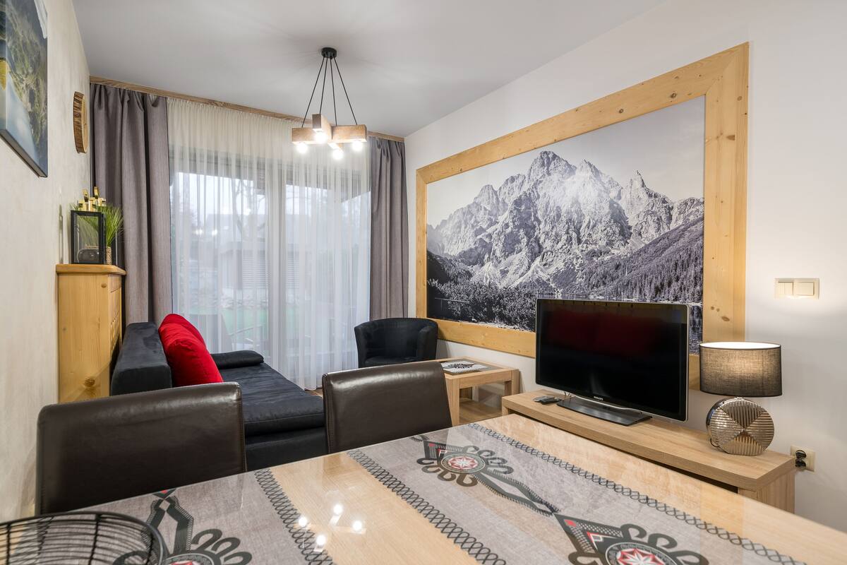 A modern living room with a sofa, a dining table, a TV, and a large mountain painting on the wall.