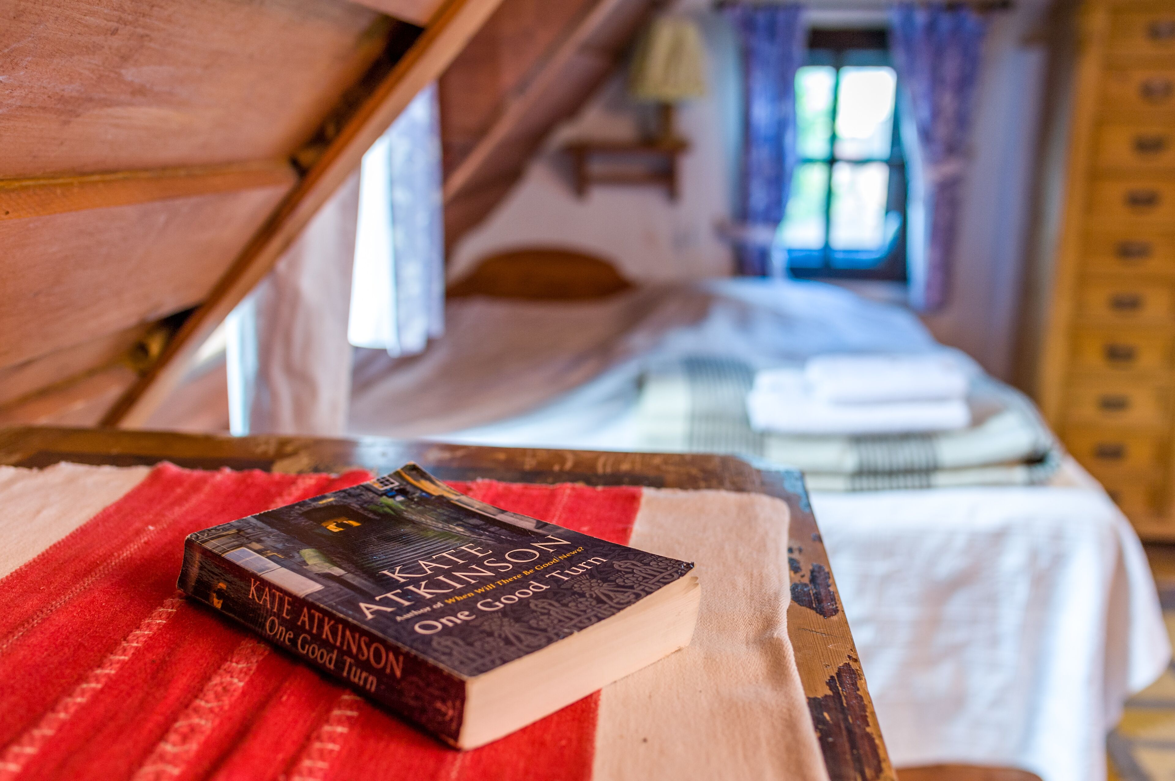 Ein Buch mit dem Titel 'One Good Turn' von Kate Atkinson auf einem Holztisch in einem Zimmer mit Bett und Fenster.