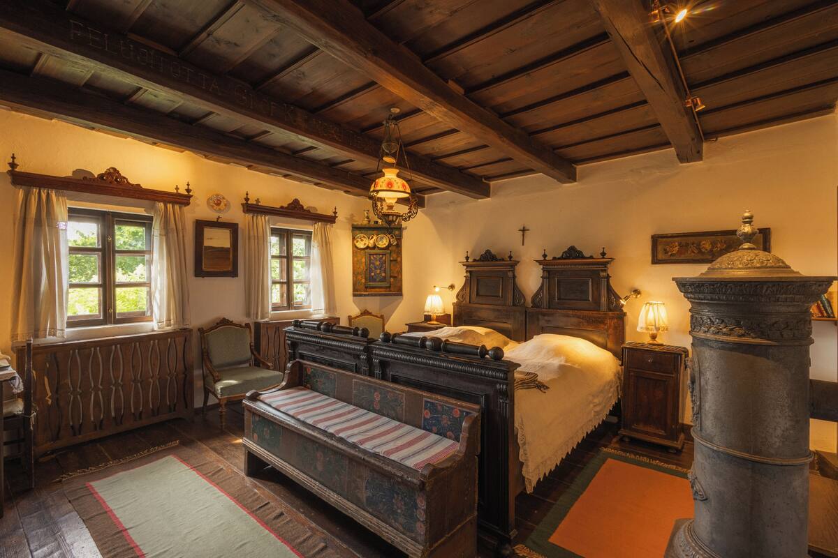 Une chambre traditionnelle avec un lit, un banc, une malle en bois et une grande colonne.