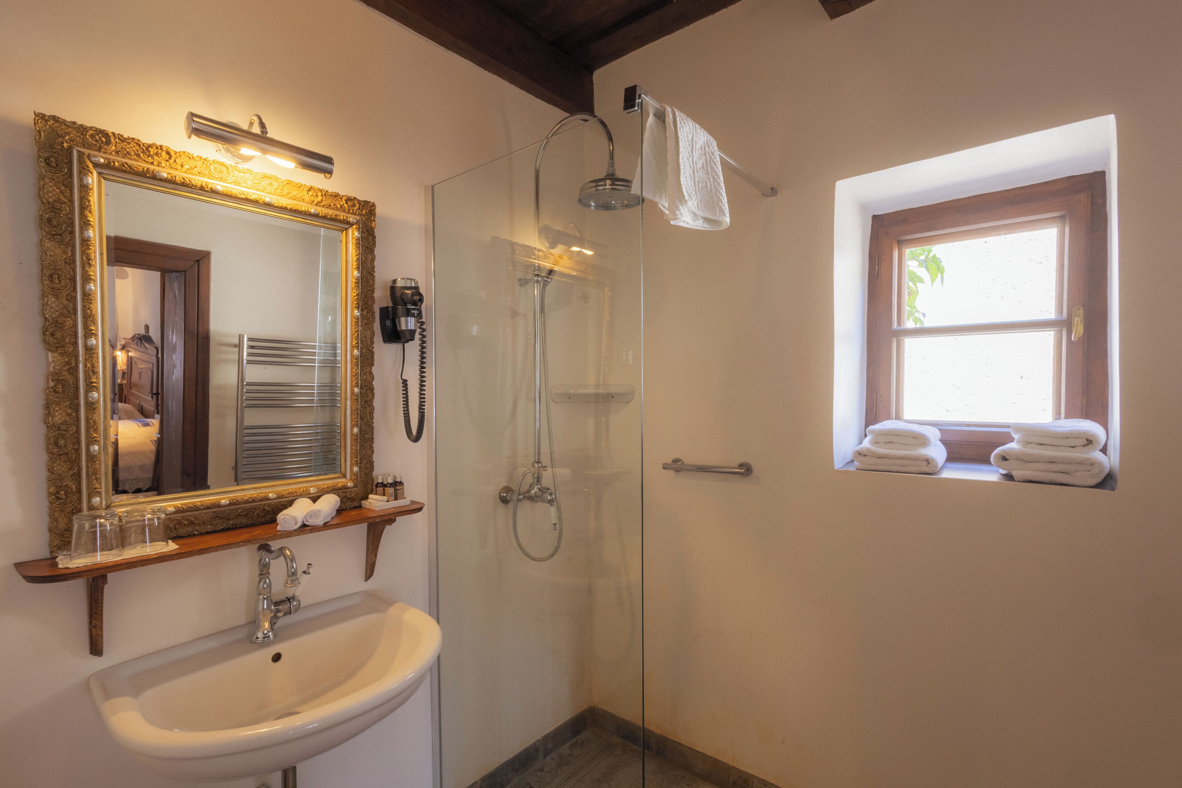 A bathroom with a sink, mirror, shower, and a window with towels.