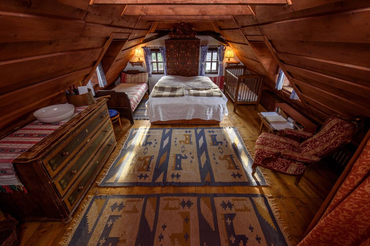 Une chambre confortable située dans les combles, avec un plafond en bois, un grand lit, une commode, une chaise et une fenêtre avec des rideaux.