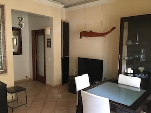 Ein Essbereich mit einem Glastisch, weißen Stühlen, einem Fernseher und einem dekorativen Holzelement an der Wand.