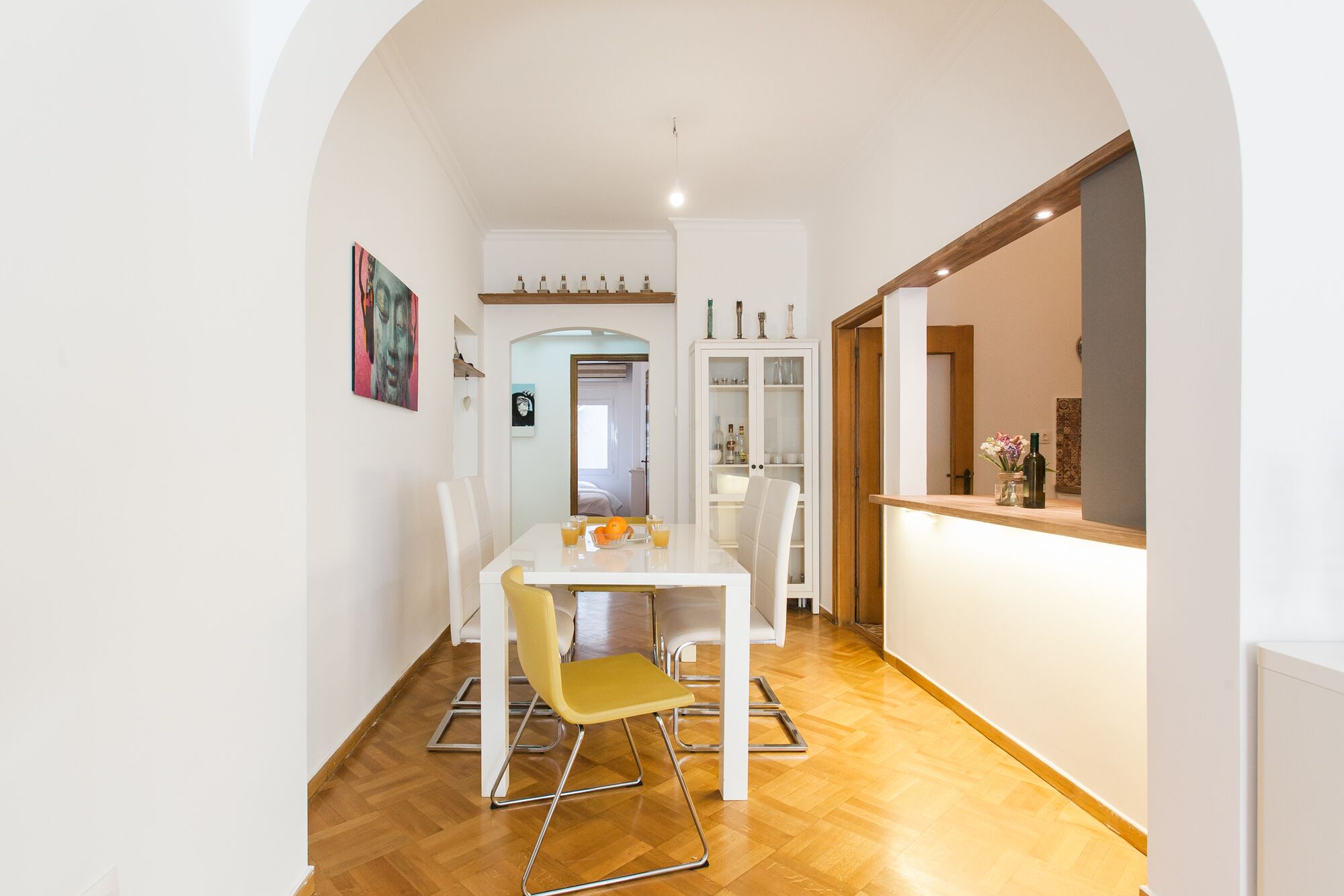 Un comedor con una mesa blanca y sillas amarillas, un armario con puertas de cristal y suelo de madera.