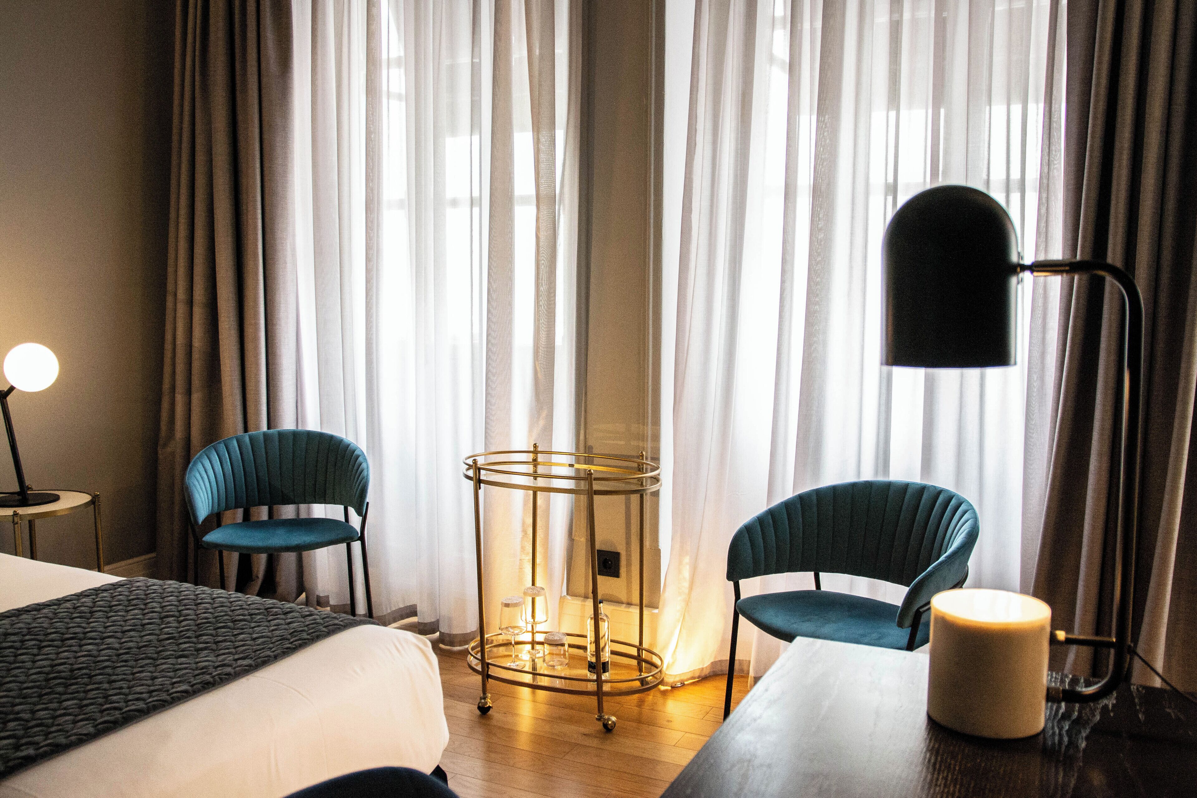 A hotel room with a bed, two chairs, a table, a lamp, and a side table with a candle.