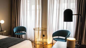 A hotel room with a bed, two chairs, a table, a lamp, and a side table with a candle.