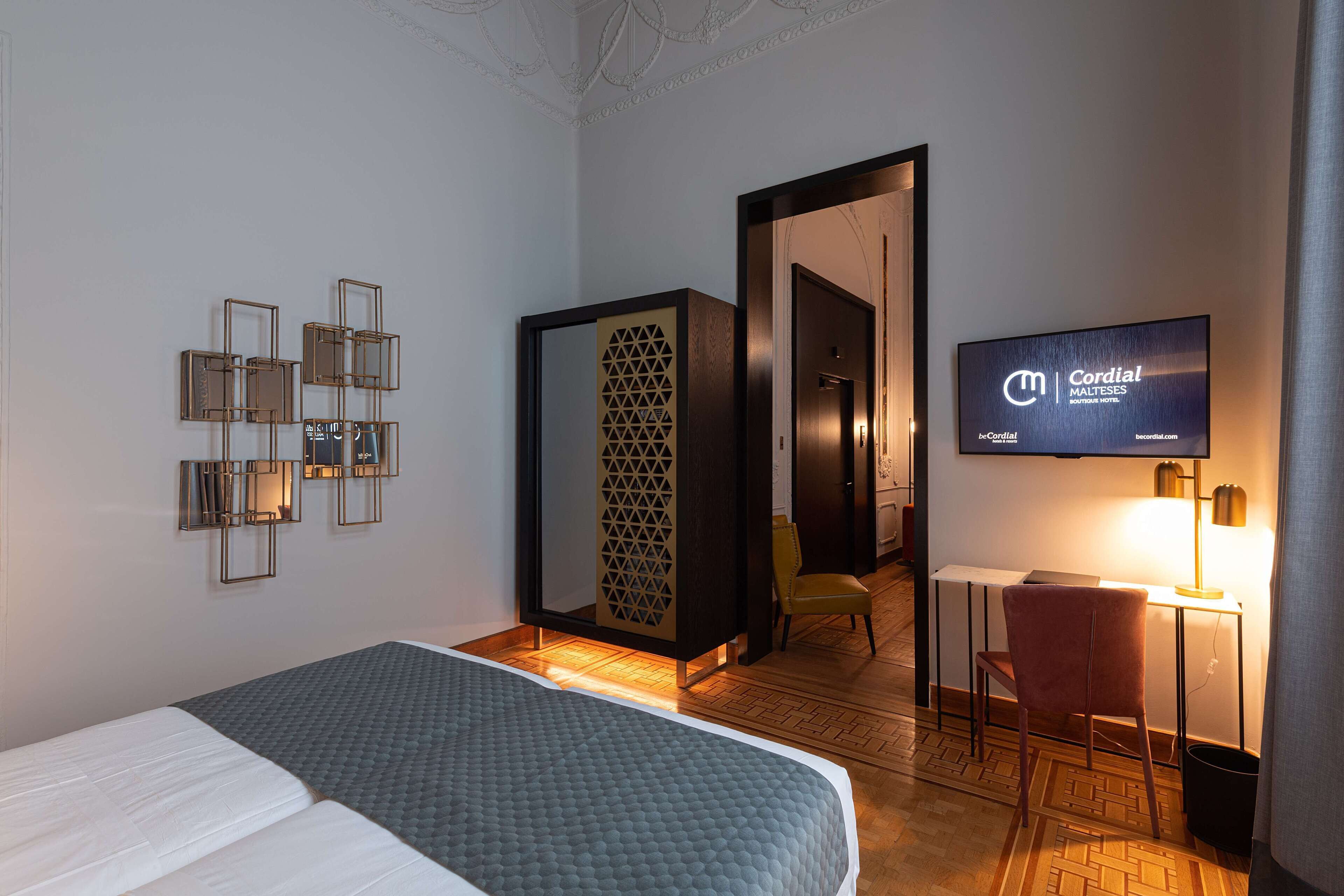 A hotel room with a bed, a desk with a chair, a TV, and a decorative wall panel.