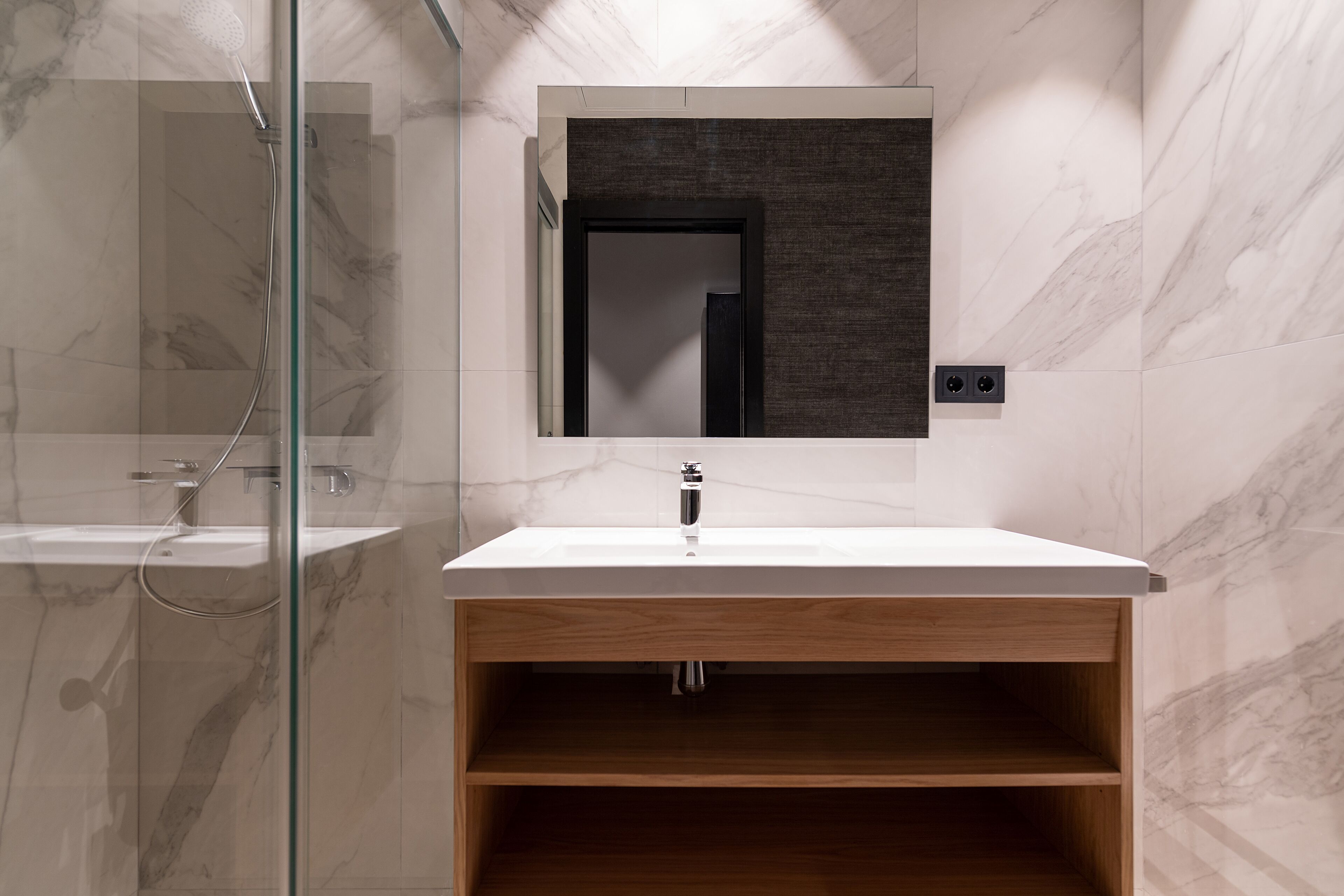 A modern bathroom with a glass shower enclosure, a wooden vanity with a mirror, and a marble-tiled wall.