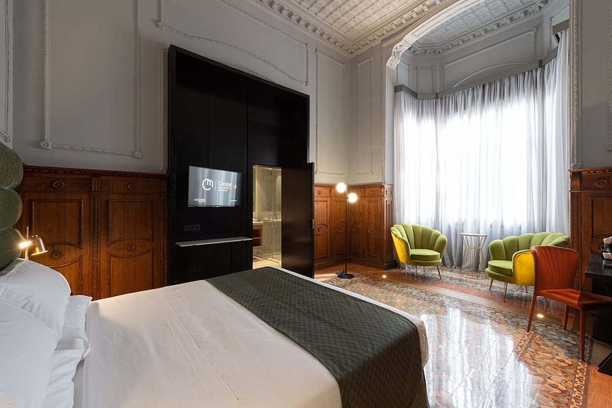 Une chambre d’hôtel avec un grand lit, une télévision, des panneaux de bois au mur, des fauteuils verts et jaunes, et une grande fenêtre avec des rideaux.
