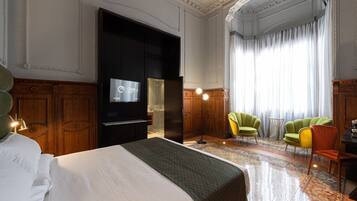 Una habitación de hotel con una cama grande, un televisor, paneles de madera, sillas verdes y amarillas, y un ventanal con cortinas.