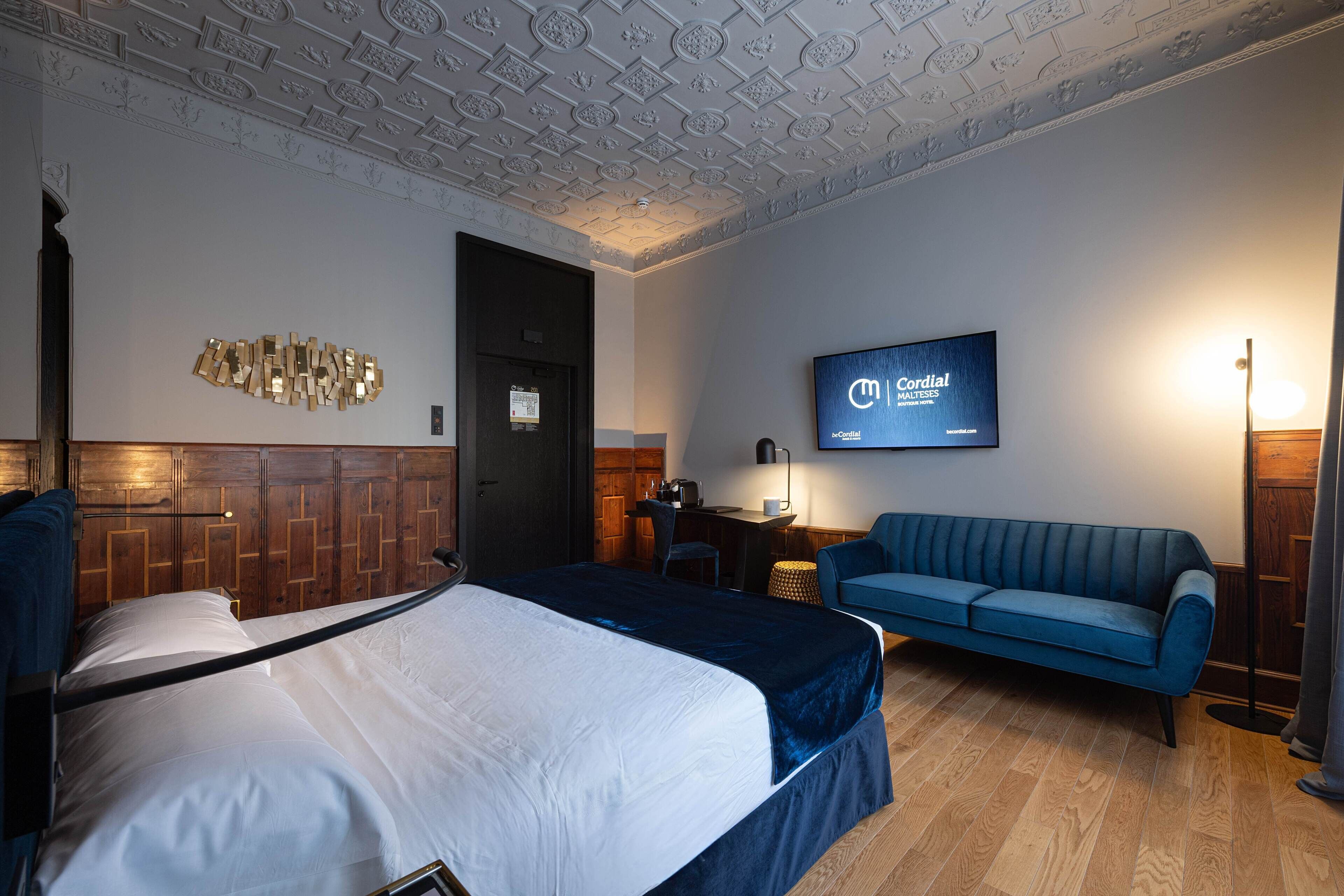 A hotel room with a bed, a blue sofa, a desk with a chair, a television mounted on the wall, and a wooden panel wall design.
