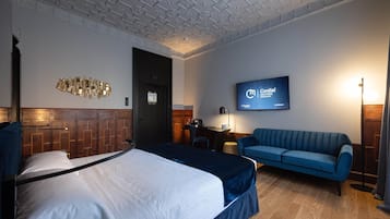 A hotel room with a bed, a blue sofa, a desk with a chair, a television mounted on the wall, and a wooden panel wall design.