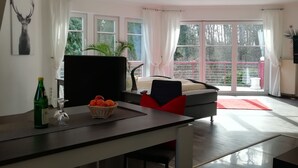 A modern room with a bed, a desk with a basket of fruit, a bottle, and a glass. There are windows with curtains, a balcony with a railing, and a plant. - Comfort apartment in the wildlife park (Koblenz)