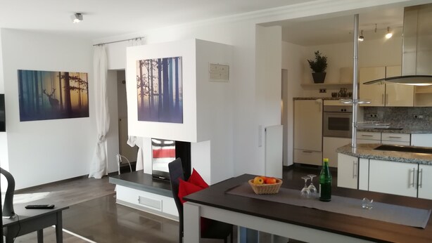 A modern kitchen with white cabinets, a built-in oven, and a granite countertop. There is a dining area with a table and chairs, and a painting of a forest scene on the wall. - Comfort apartment in the wildlife park (Koblenz)
