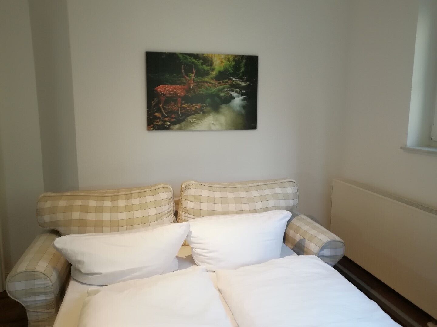A room with a bed, two checkered armchairs, a painting of a deer and waterfall on the wall, and a window on the right.