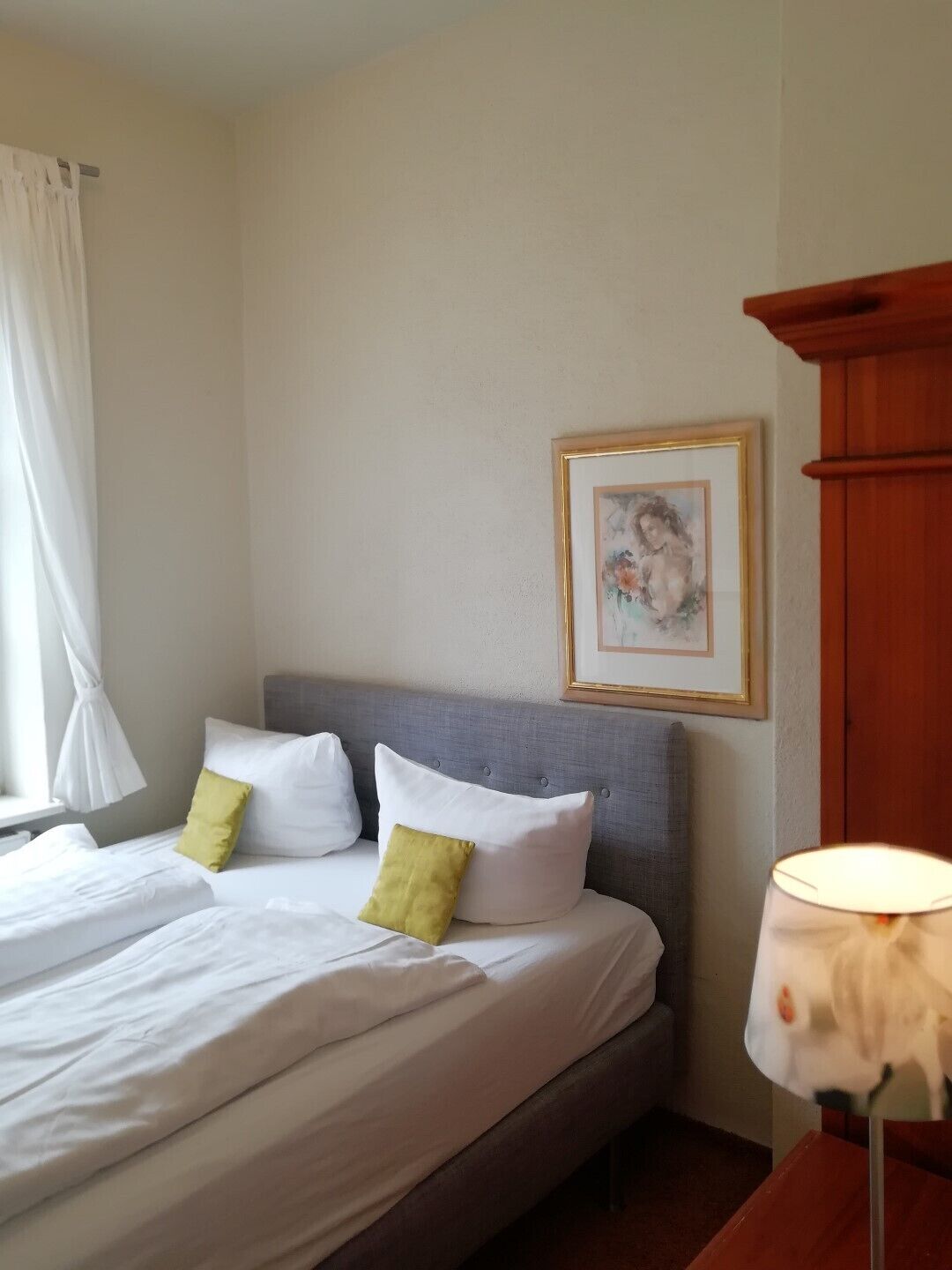 A neatly made bed with white bedding and two pillows, one green and one white, in a room with a grey upholstered headboard. A framed picture hangs on the wall above the bed.