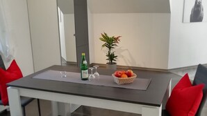 Un espace repas moderne avec une table, des chaises et un banc. On peut y voir une petite plante sur la table, une bouteille d’eau et un panier de fruits.