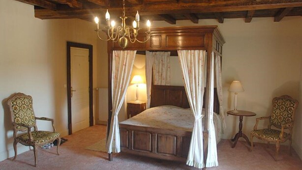 A four-poster bed with a wooden frame and white curtains, flanked by two upholstered chairs with patterned upholstery, in a room with a wooden ceiling and a small round table with a lamp. - Four poster bedroom (launay blot)