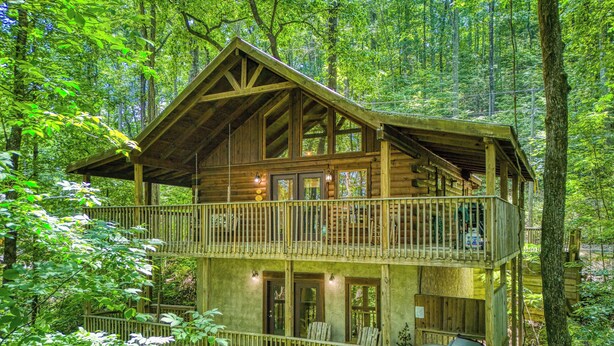 A two-story wooden cabin with a balcony, surrounded by dense forest.
