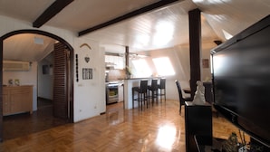 An attic-style room with wooden floors, a dining area with chairs, a kitchen with appliances, and a large black TV. - The Grich (Zagreb)