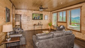 A log cabin interior with a fireplace, leather sofas, a dining table, and a large window offering a view of the outdoors.