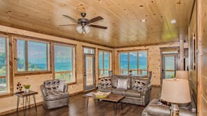 A spacious living room with wooden walls and floors, large windows, a ceiling fan, and a sofa set.