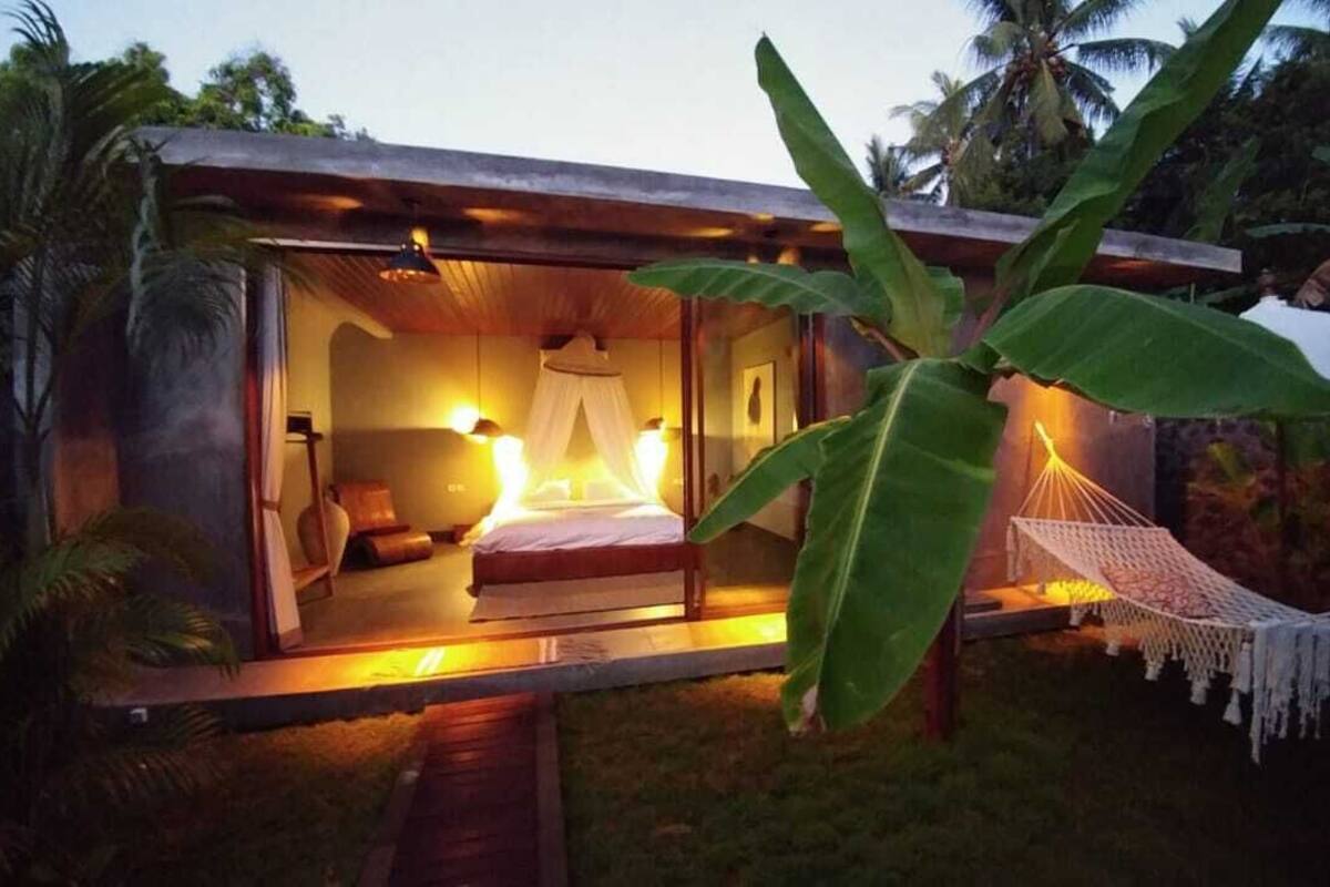 Quarto aconchegante e bem iluminado, com cama, mesa de cabeceira e uma rede lá fora.